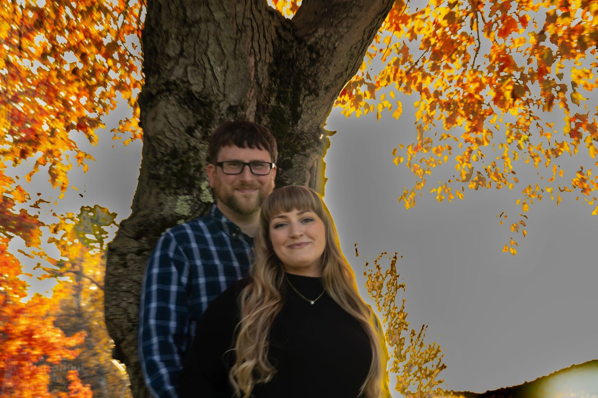 A couple stands closely together in front of a large tree with vibrant orange autumn leaves.