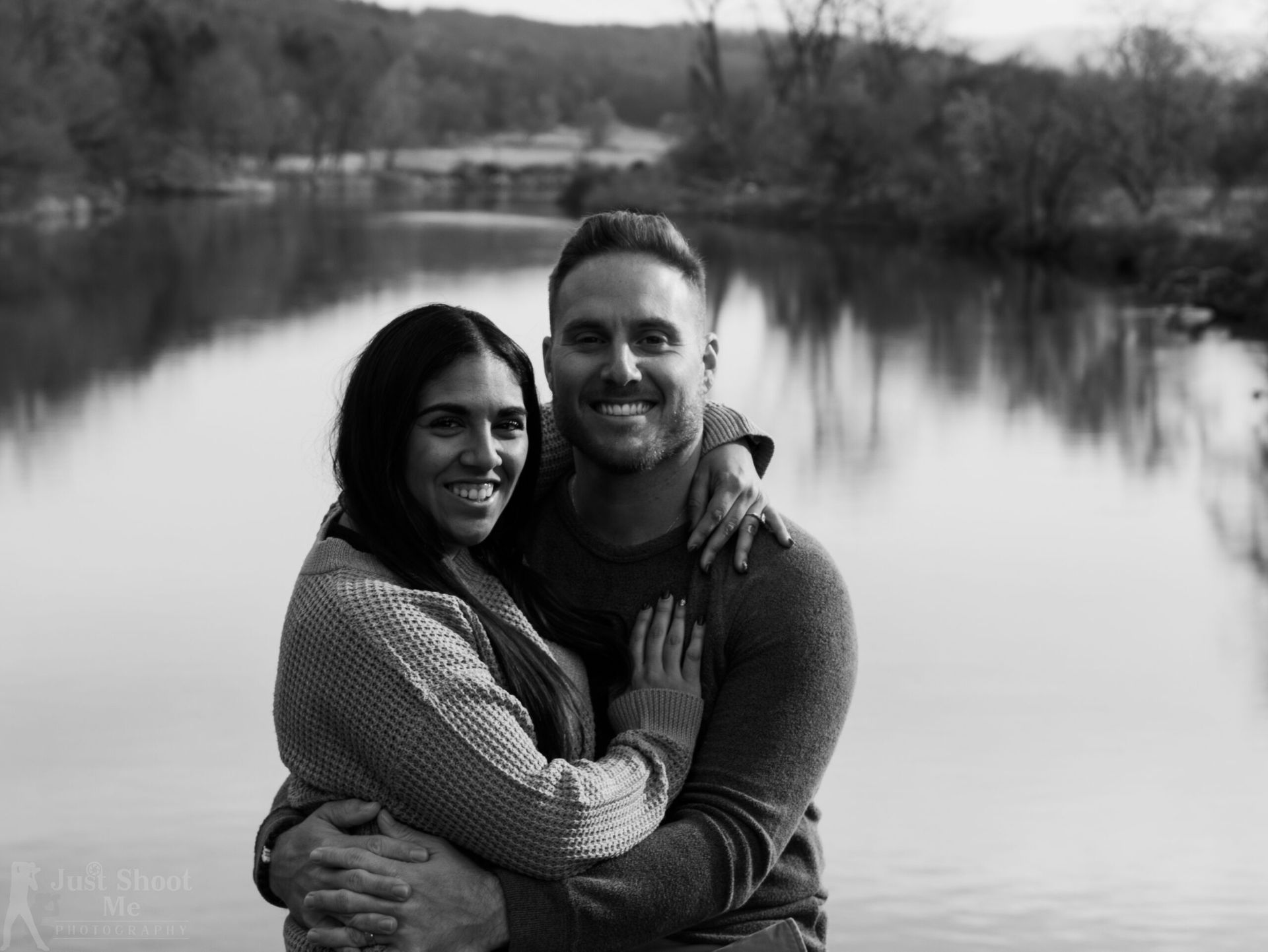 A black-and-white portrait of a couple embracing by a lake with trees in the background.