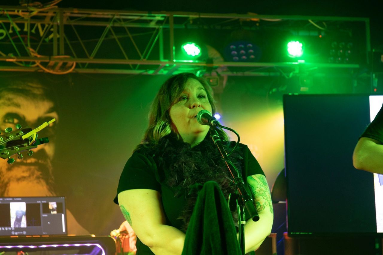 A musician sings into a microphone on a stage lit with green spotlights, with a dark background and equipment visible.