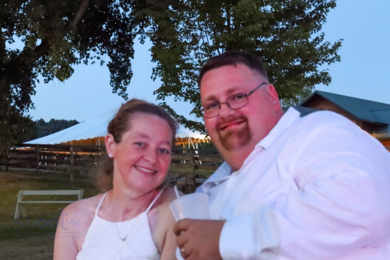 A smiling couple posing for a photo outdoors at dusk, with a tent and trees in the background.