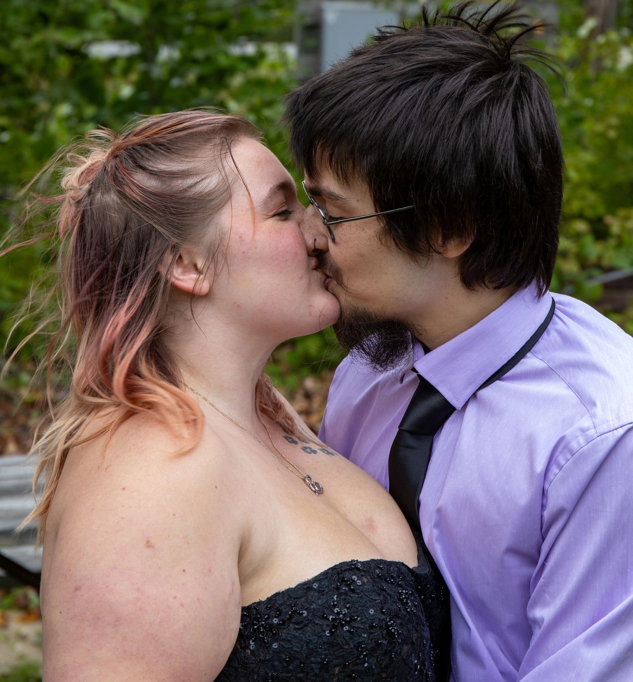 A couple sharing a kiss outdoors; one wears a black strapless dress and the other a purple dress shirt and black tie.