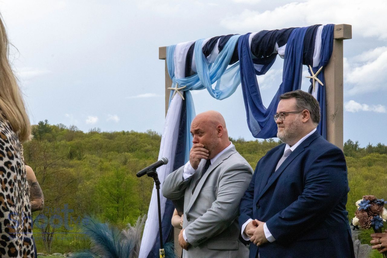 Two men in suits standing at a wedding altar draped with blue and white fabric, with one looking emotional.