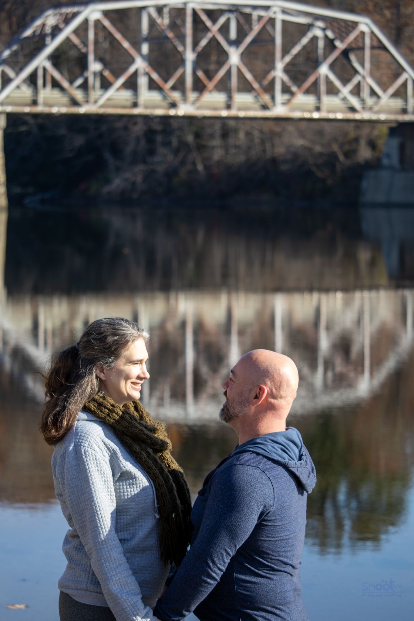 A couple in casual clothing faces each other and smiles, standing near a reflective river with a metal bridge overhead.
