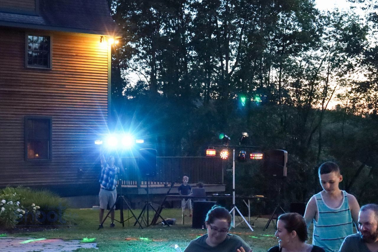 An outdoor evening party with a DJ setup, bright stage lights, and a wooden house in the background.