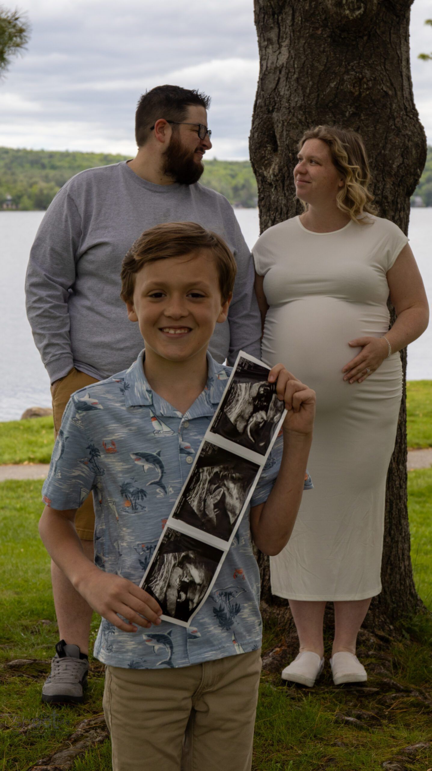 A person holds an ultrasound printout, smiling, with a couple standing behind them near a tree and a lake.