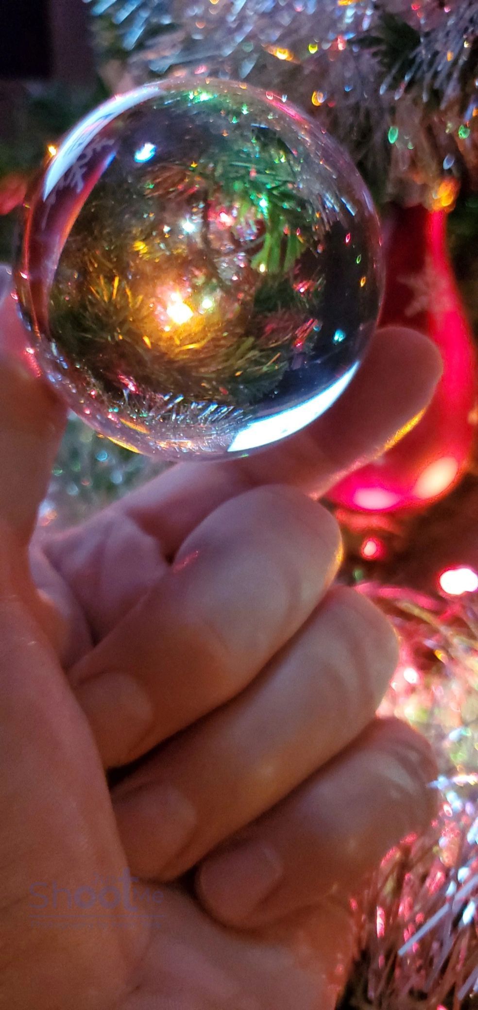 A hand holds a clear glass sphere, reflecting colorful holiday lights against a blurred Christmas tree background.