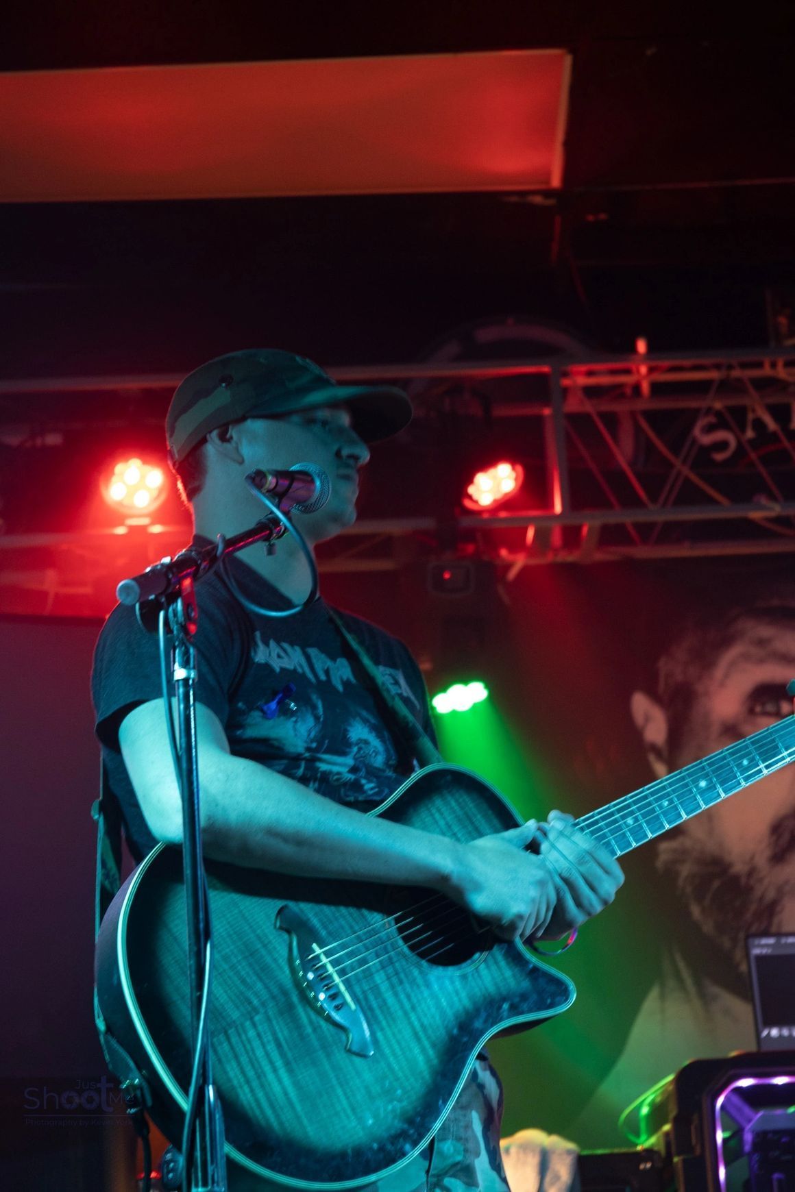 A musician in a cap playing an acoustic guitar on a dimly lit stage with red and green spotlighting.