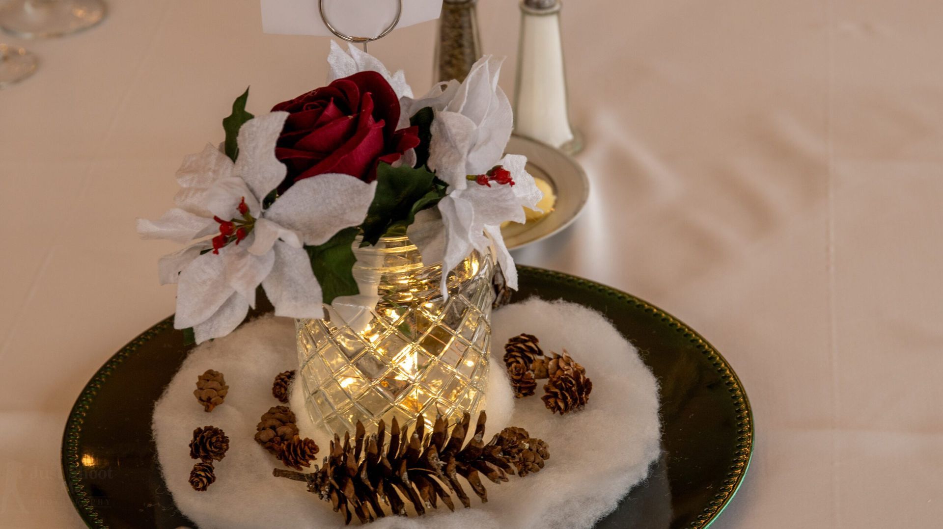 A festive centerpiece with a red rose and white poinsettias in a lit glass vase on a snowy, pinecone-decorated green plate.