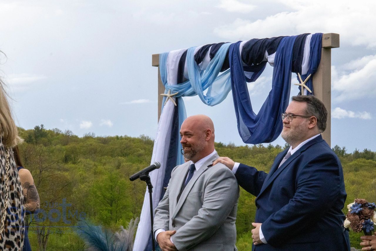 Two people in suits stand smiling under an outdoor arch decorated with blue and white fabric against a forest backdrop.