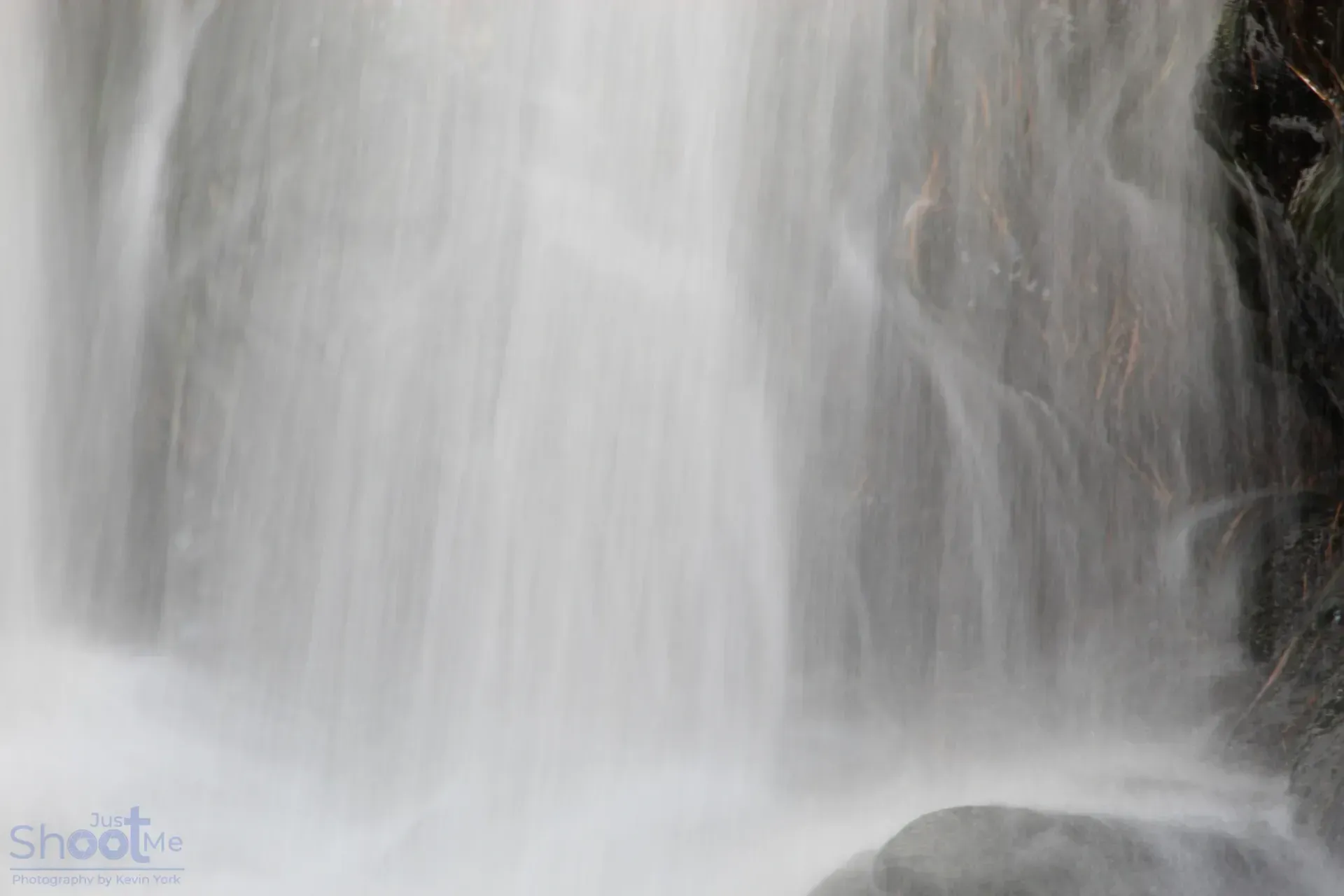 A close-up view of a waterfall with blurred, flowing white water cascading over dark, mossy rocks.