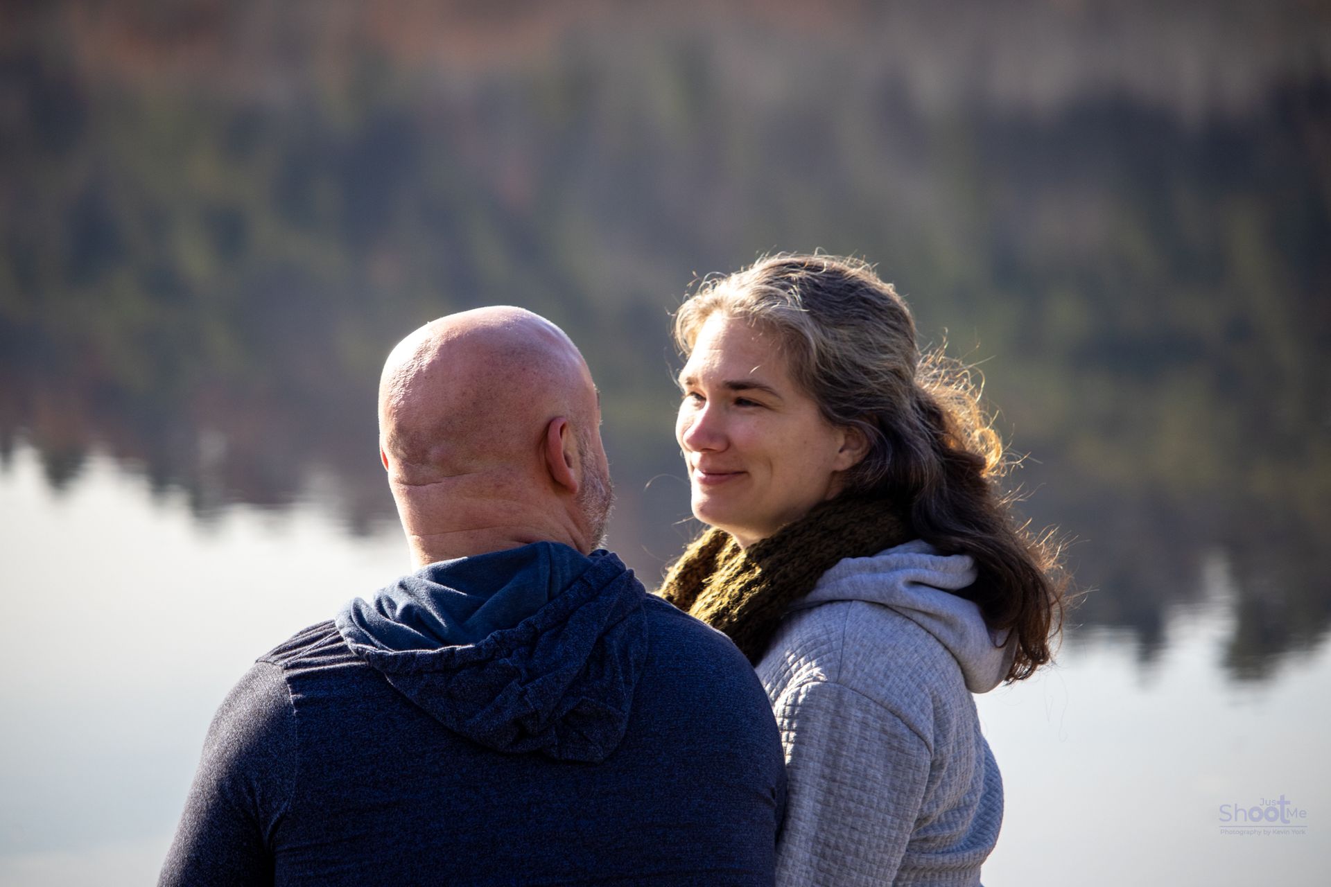 Two people stand near a calm body of water, facing each other and smiling.