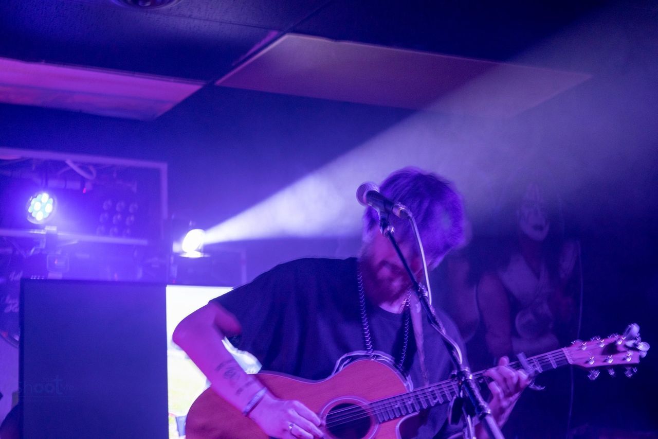 A musician plays an acoustic guitar on a dimly lit stage, illuminated by a bright beam of purple spotlight.