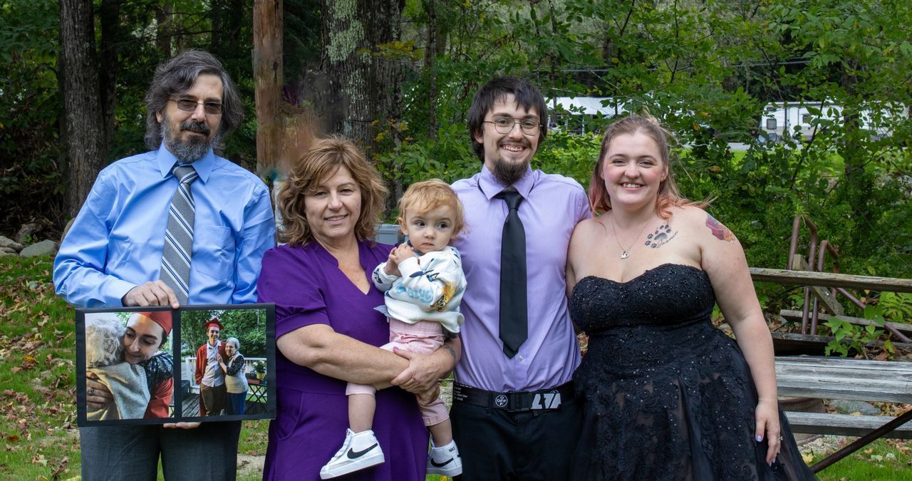 A group of four adults and a small child stand together outdoors, with one man holding a framed photo of the same group.
