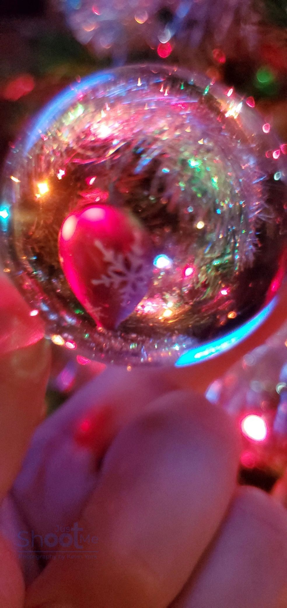A close-up of a clear glass Christmas ornament containing a small red bulb, reflecting colorful festive lights.