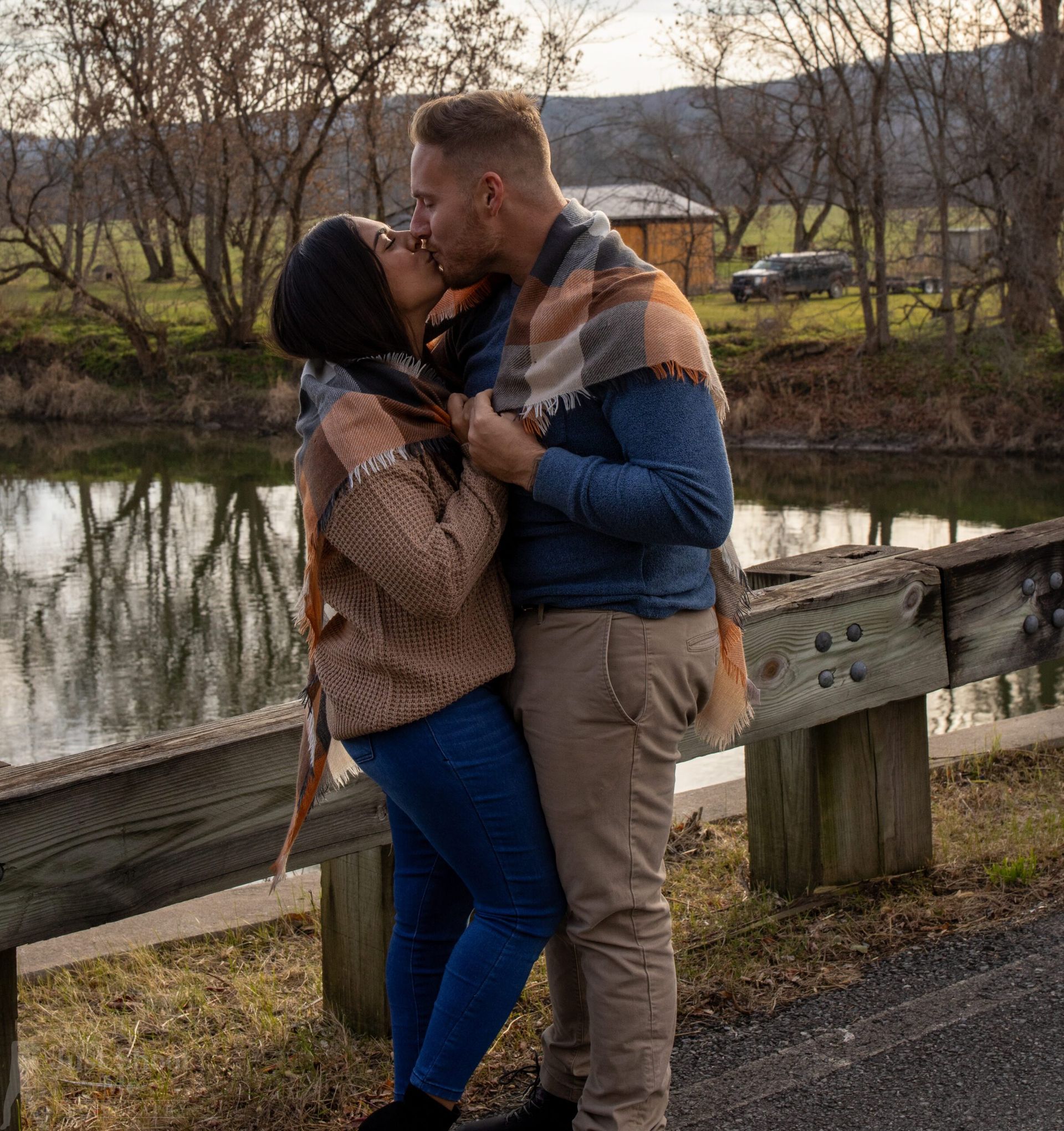 A couple shares a kiss outdoors by a lake, wrapped together in a plaid scarf, standing next to a rustic wooden bridge.