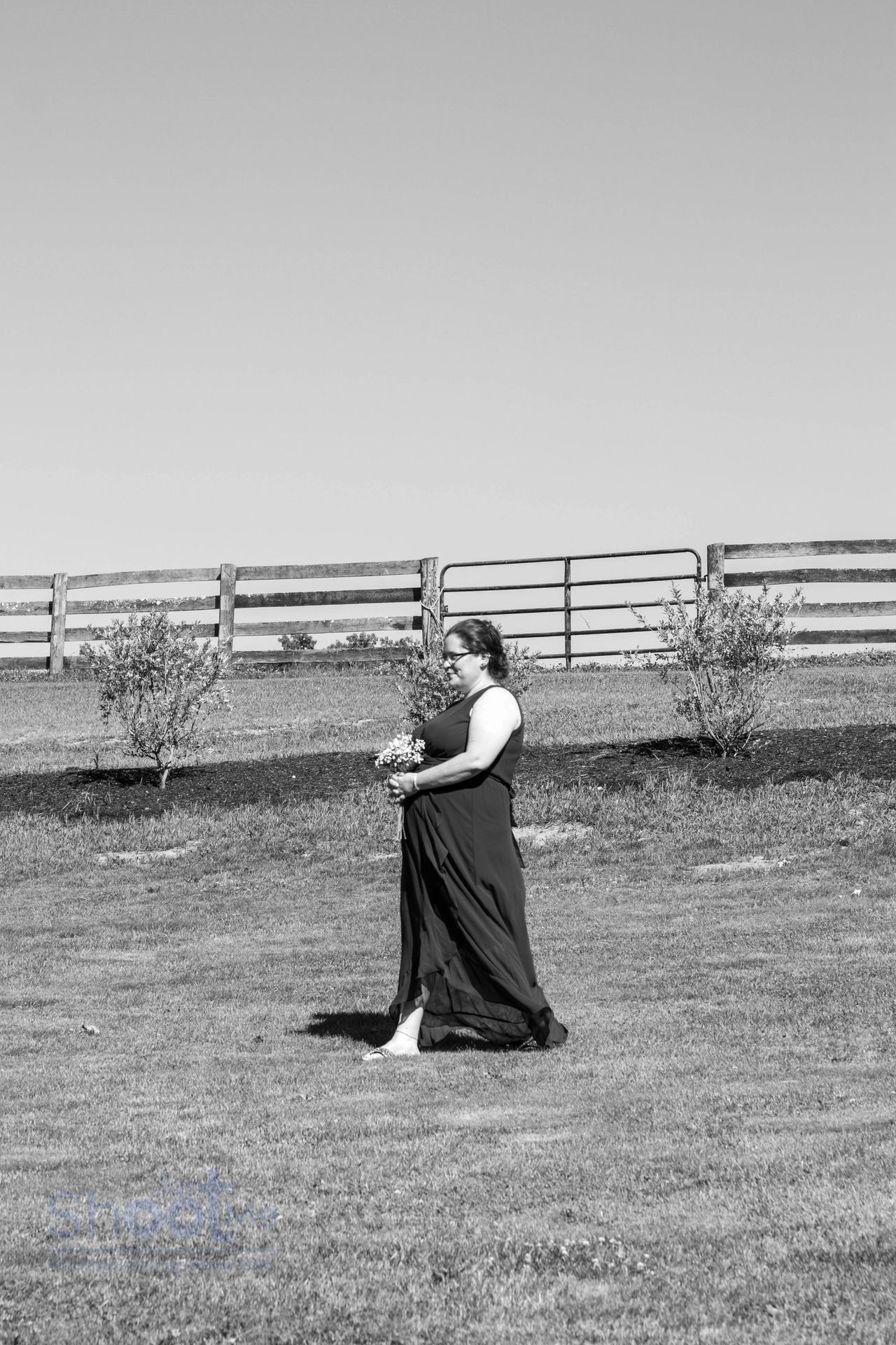 A person in a flowing dress walks across a grassy field near a split-rail fence under a clear sky.
