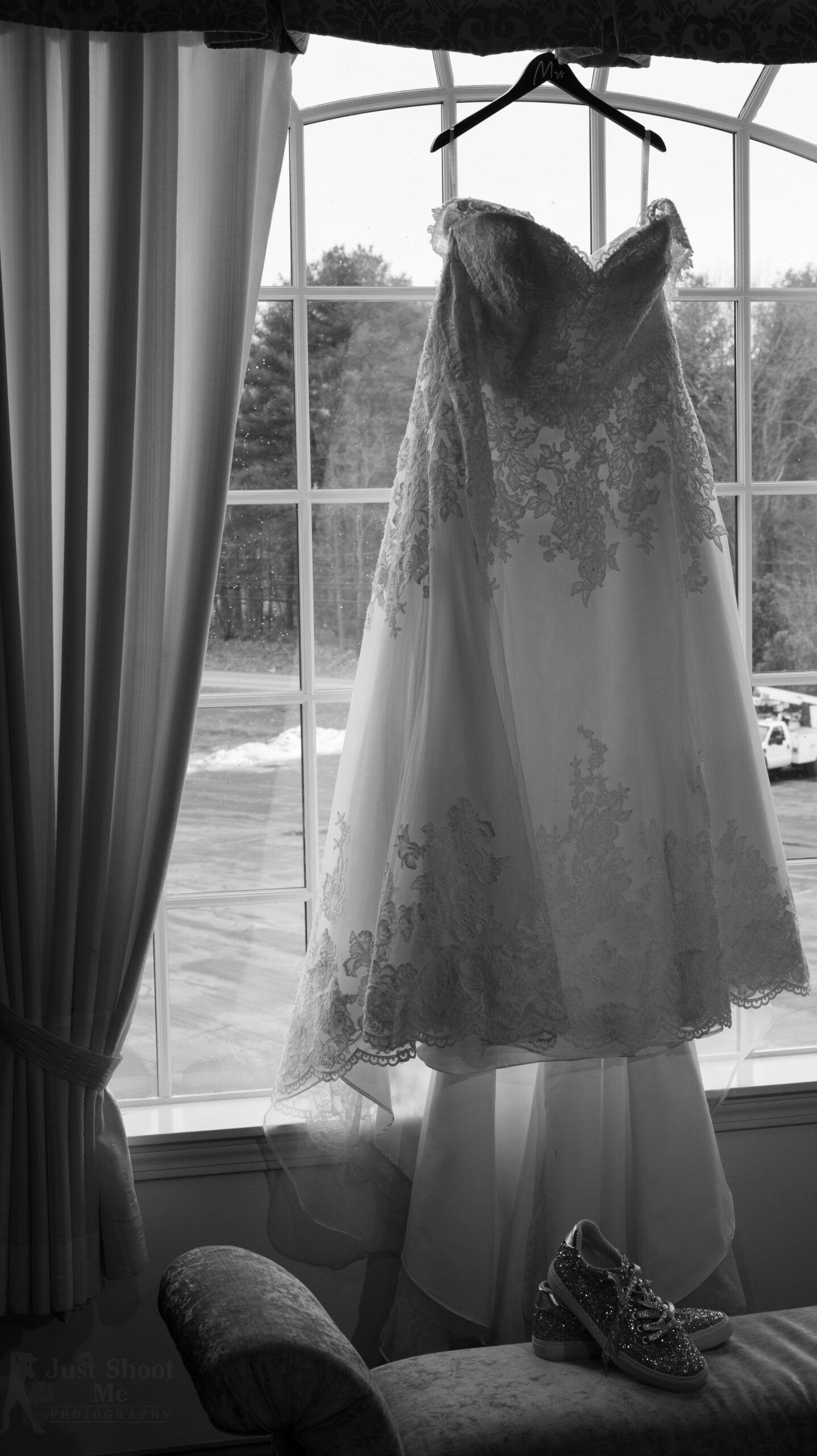 A wedding dress hangs in front of a window, with a pair of embellished heels resting on a bench below.