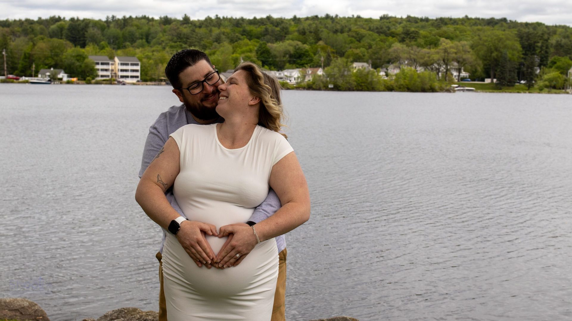A pregnant person in a white dress and their partner pose by a lake, forming a heart shape with their hands over the belly.