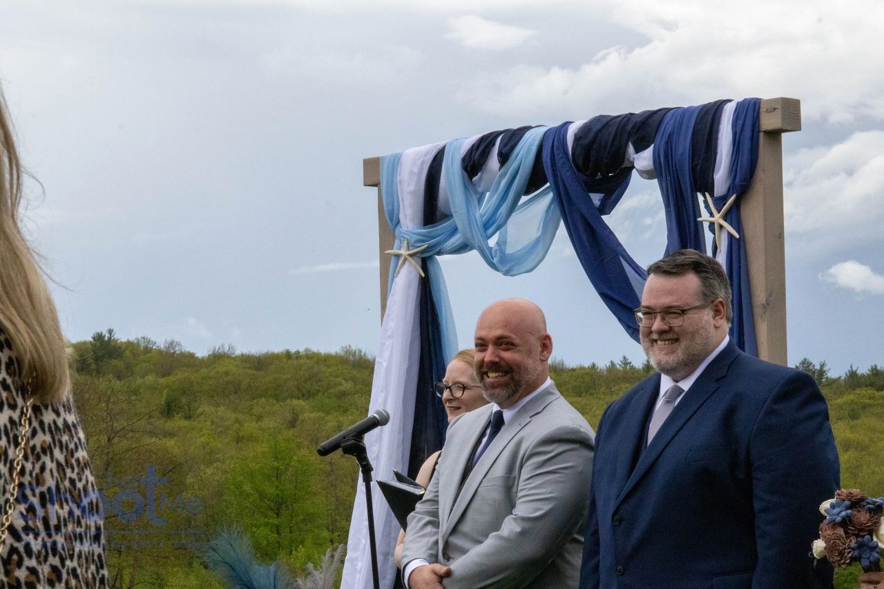 Two smiling people in suits stand before an outdoor wedding arch draped in blue and white fabric.