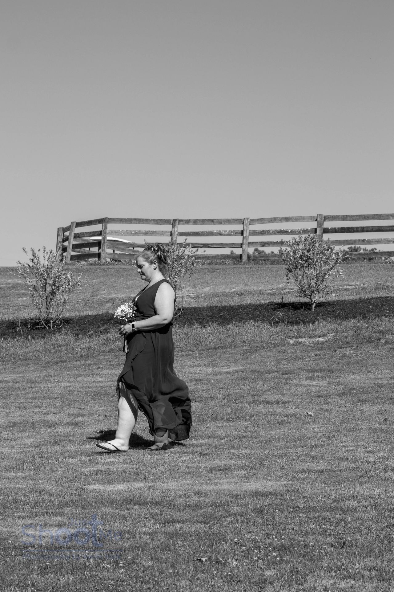 A person in a formal, dark gown holds a bouquet while walking across a grassy field near a wooden fence.