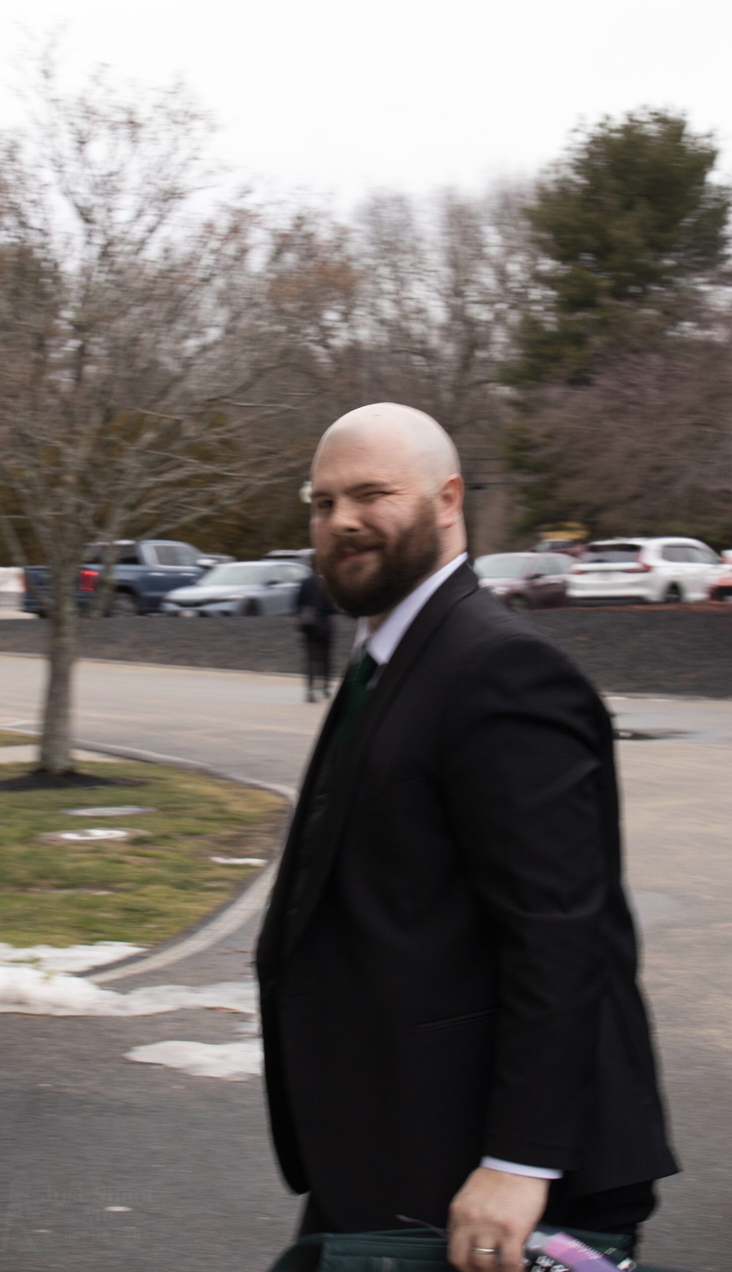 A person with a beard, wearing a black suit and tie, looking toward the camera in an outdoor, paved parking lot.