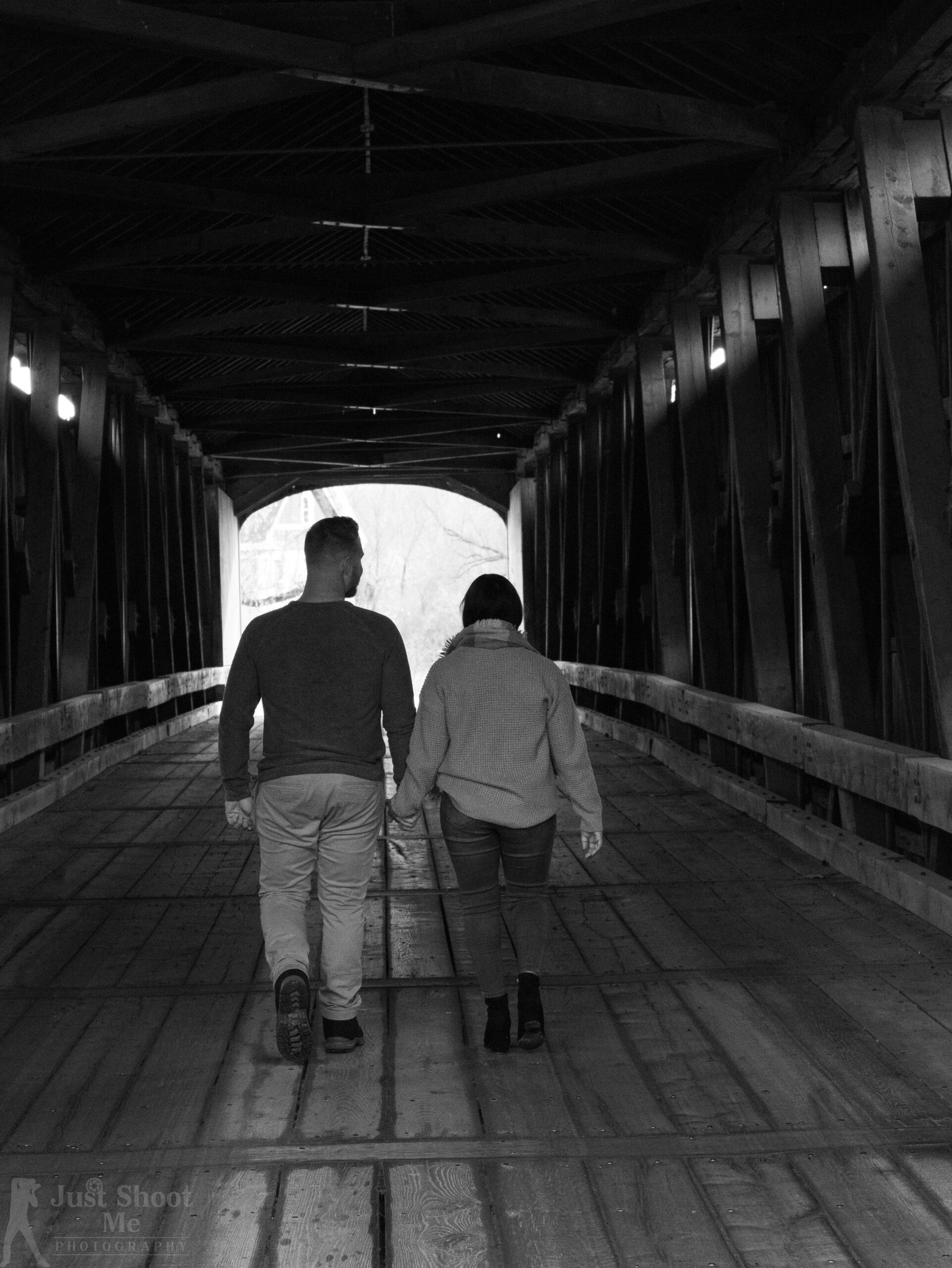A couple walks hand-in-hand through the center of a rustic, wooden covered bridge toward a bright opening.