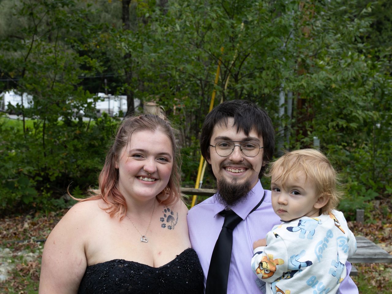 Three people pose together outdoors in front of a tree-lined background. A woman and a man hold a toddler.