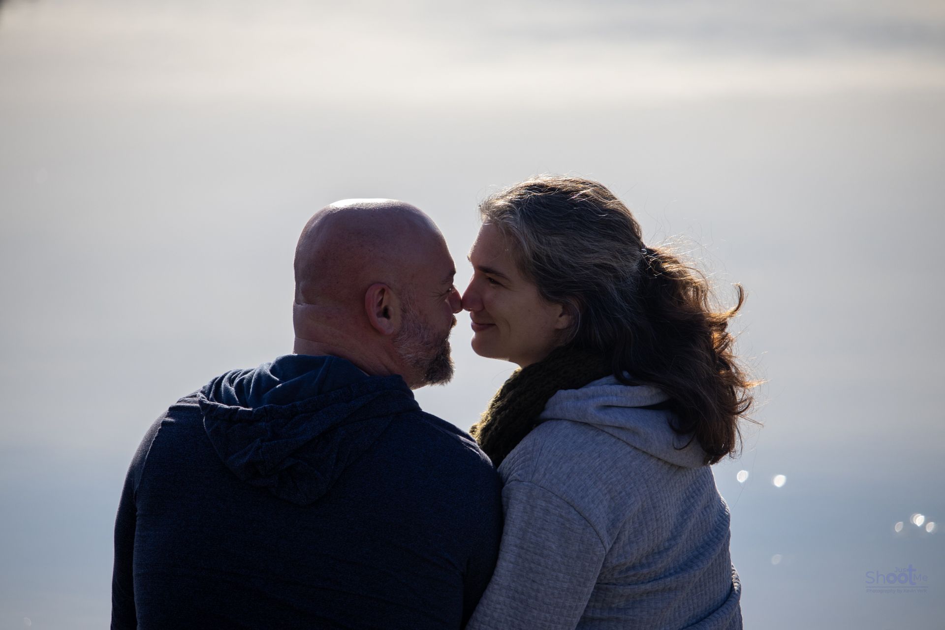 A couple face each other, noses touching, with an out-of-focus, bright sky in the background.