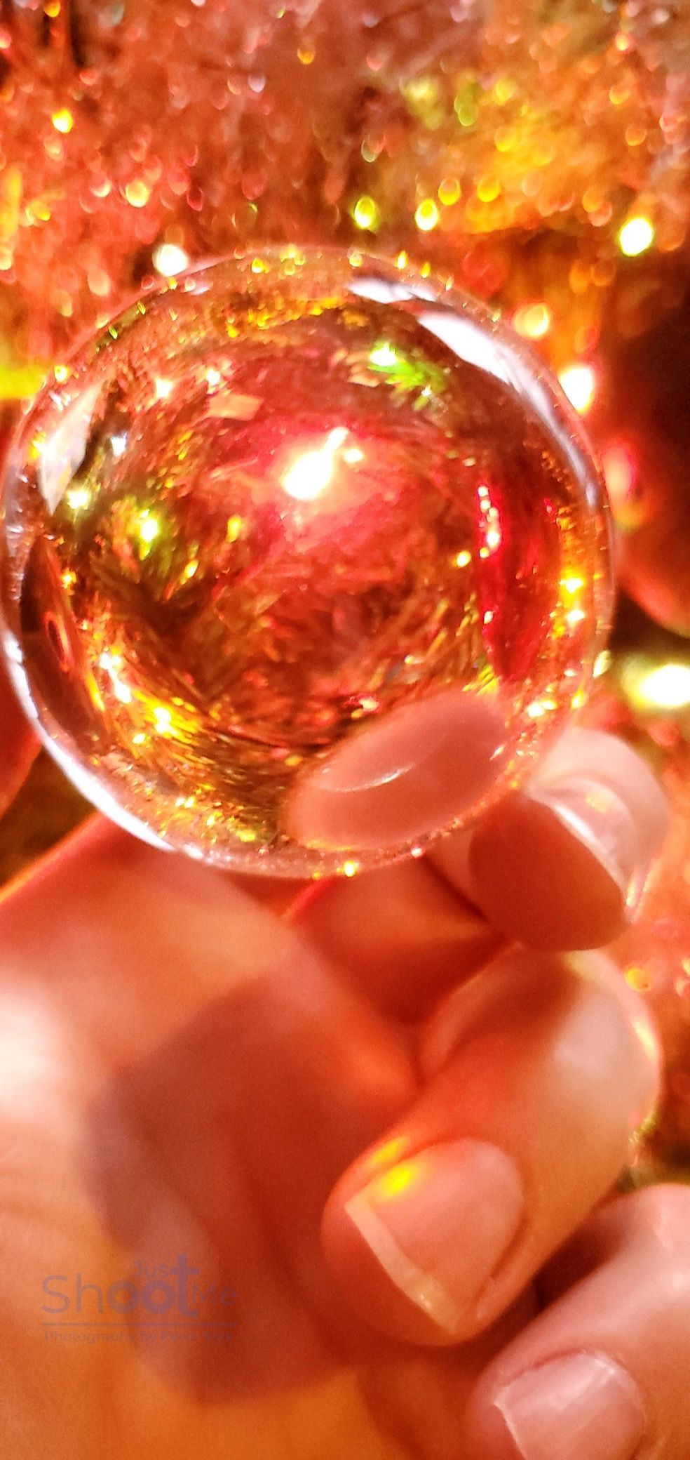 A hand holds a clear, spherical ornament filled with warm, glowing gold and red lights against a bokeh background.