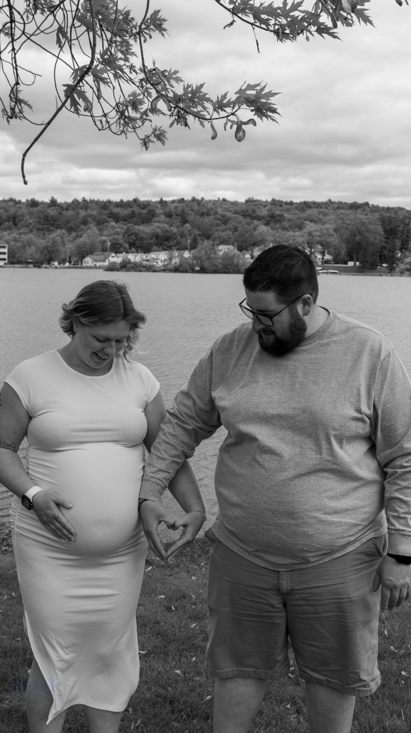 A pregnant person and their partner stand by a lake, holding hands and forming a heart shape over the baby bump.
