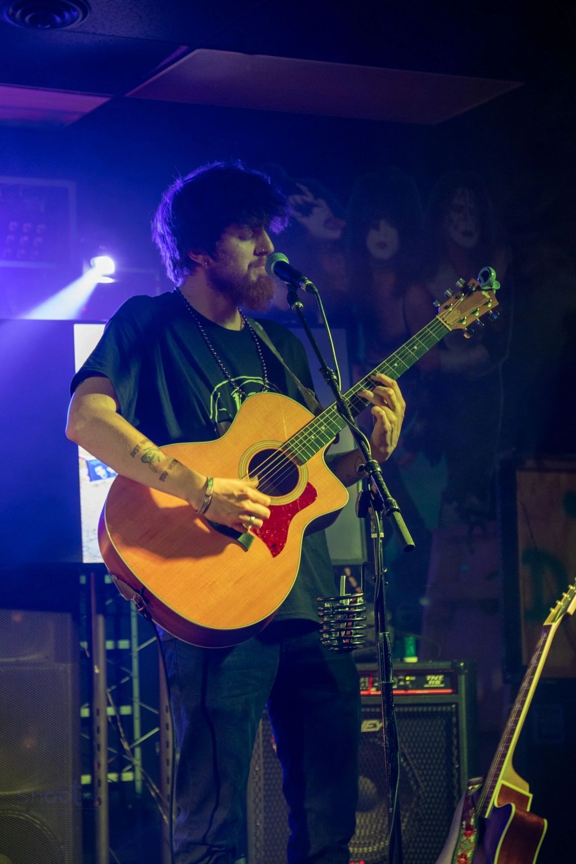 A person with a beard plays an acoustic guitar and sings into a microphone on a dimly lit, blue-hued stage.