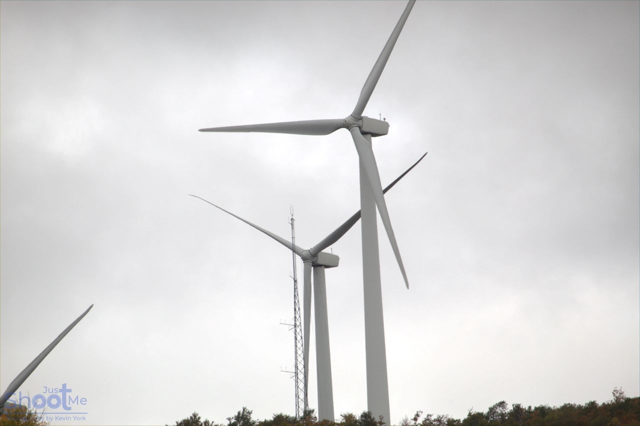 Large wind turbines stand on a hill under an overcast, gray sky, with a telecommunications tower visible between them.