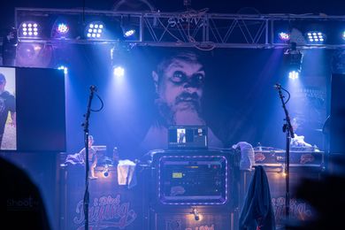 A stage with blue lighting and a large projected face behind a DJ booth, with microphones on stands and concert equipment.