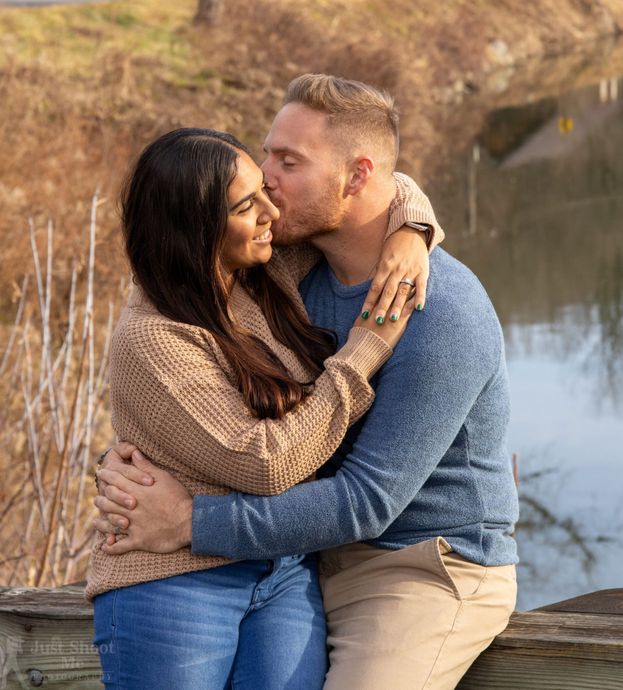 A couple embraces on a wooden railing near water, with the person on the right kissing the other on the cheek.