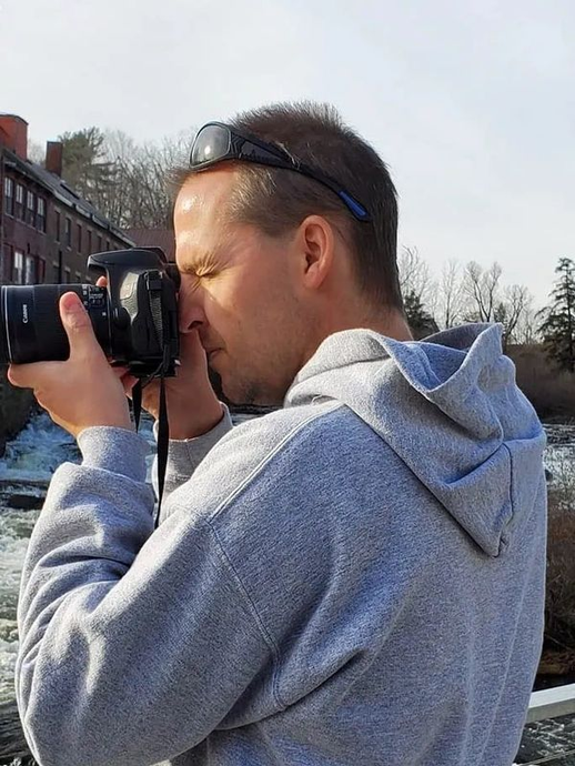 A person wearing a grey hoodie and sunglasses on their head takes a photograph with a digital camera outdoors.