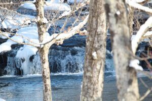 A small, flowing waterfall framed by snow-dusted trees in a winter landscape.