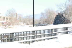A snow-covered wooden railing and benches in a park setting with trees and distant hills on a bright winter day.
