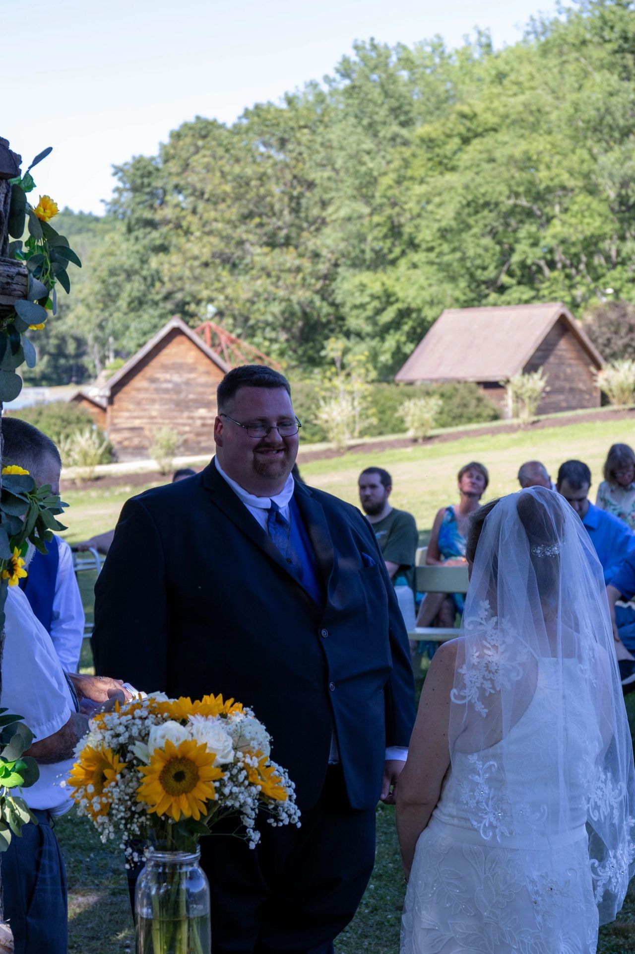 A couple stands at an outdoor wedding ceremony, facing each other with guests seated behind them in a grassy, rural area.