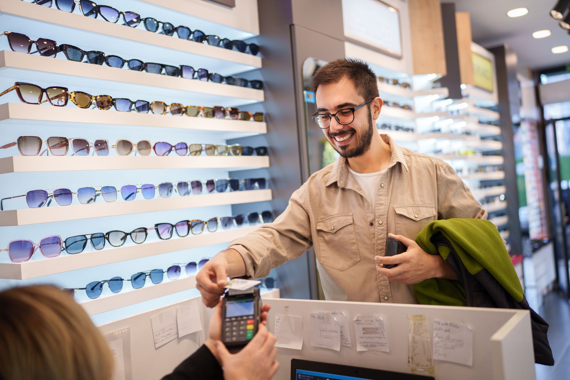 Man paying with card at an eyewear store, reaching towards a card reader near a shelf of glasses.