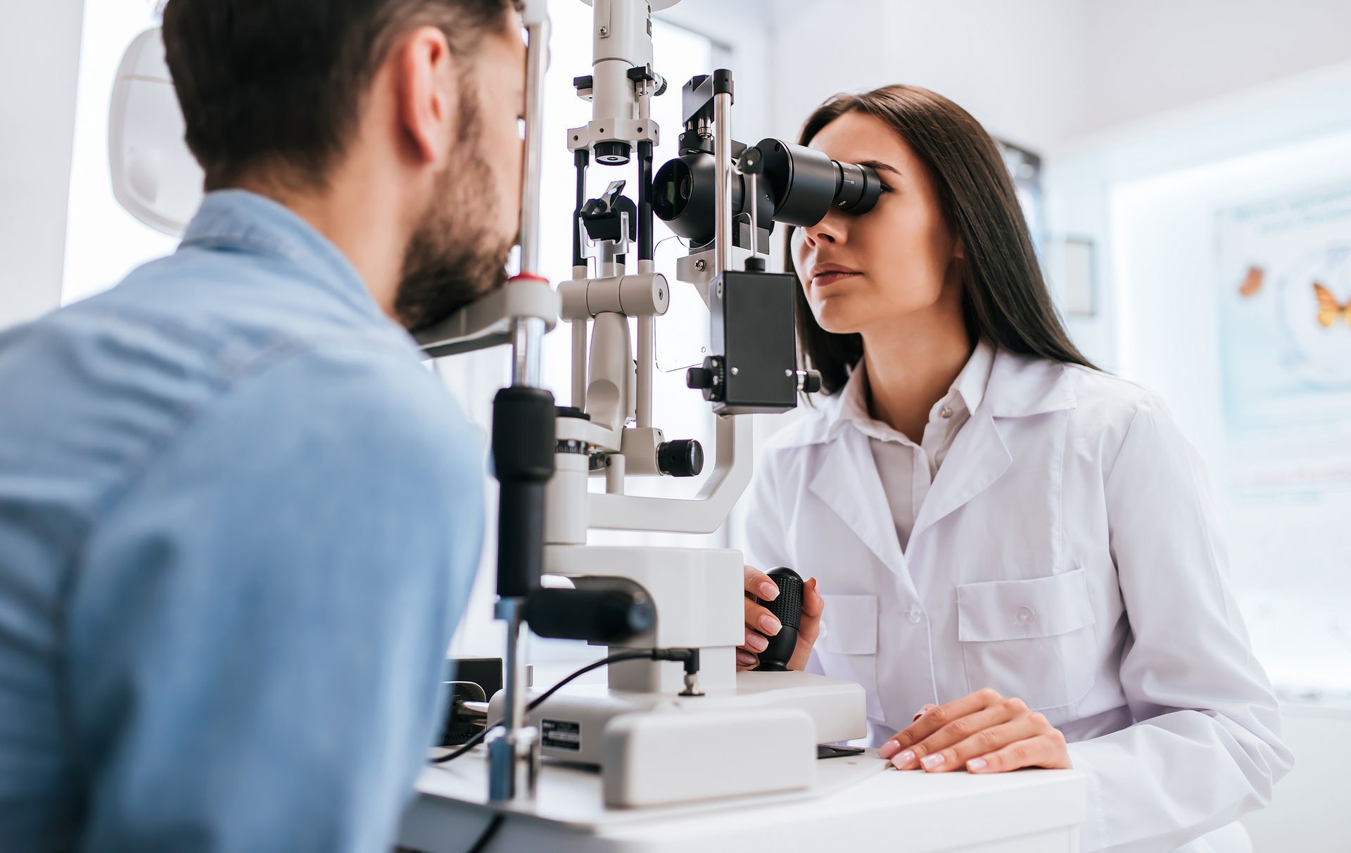 Optometrist examining a patient's eyes with a slit lamp in an exam room.
