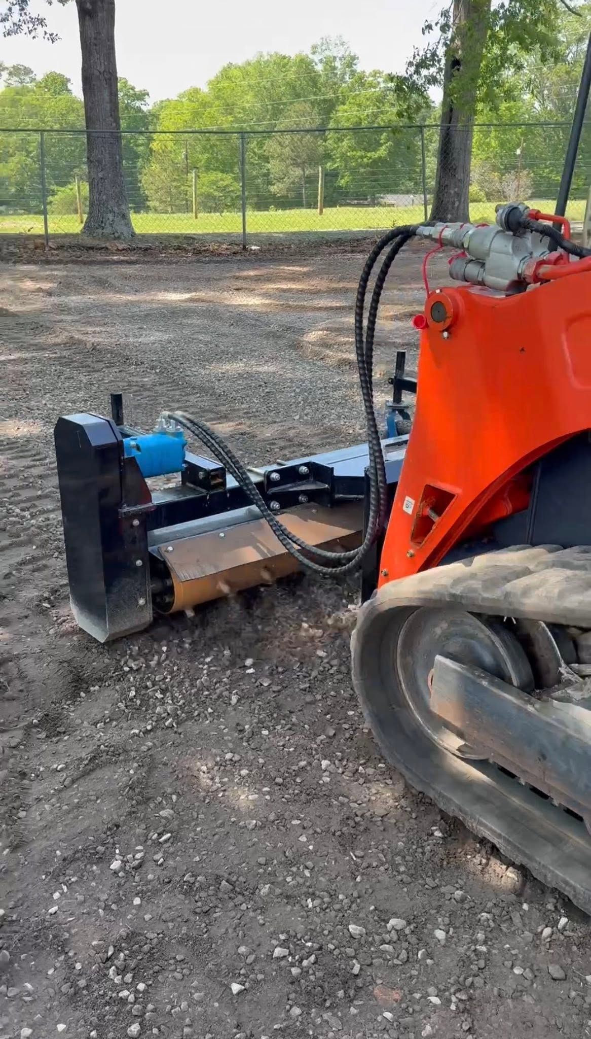 Orange skid steer with a brush cutter attachment clearing dirt in a wooded area.