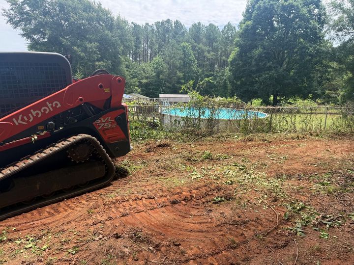 Red skid-steer loader on a dirt lot near a fenced pool, with trees in the background.