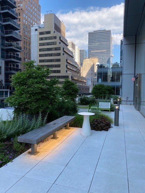 Rooftop garden with bench and white table, buildings in background. Cloudy sky, neutral tones, greenery.