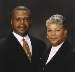 A formal portrait of two people posing side-by-side against a dark, solid background, both wearing dark professional jackets.