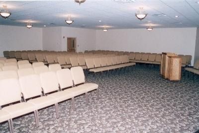 A large, empty conference room with rows of beige chairs facing a wooden podium on a patterned grey carpet.