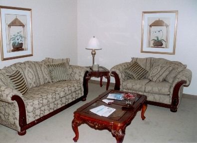 A sitting area with two beige patterned couches, a wooden coffee table, a small side table with a lamp, and wall art.