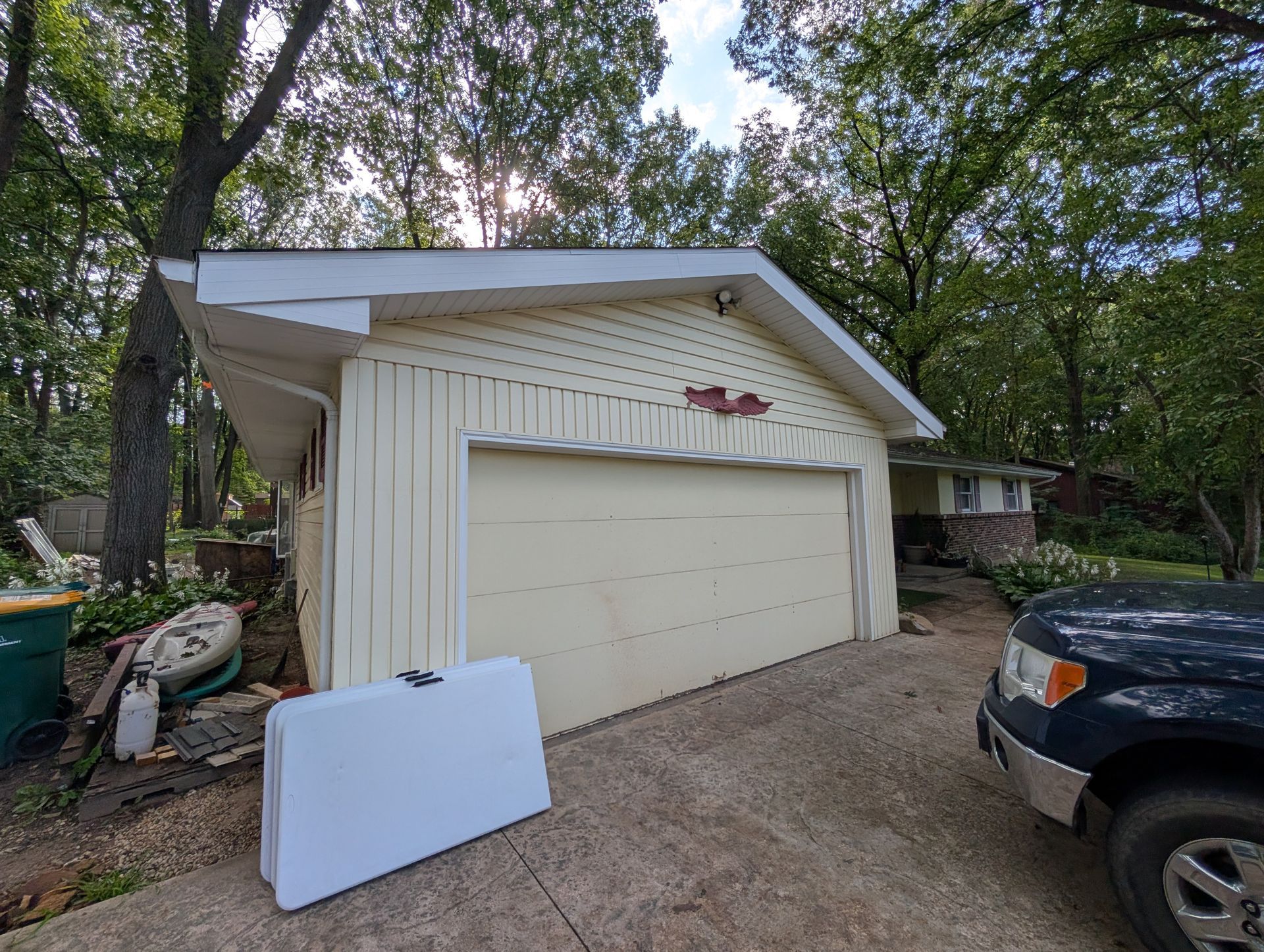 Brown ranch-style house with attached garage, surrounded by trees and a driveway on an overcast day.