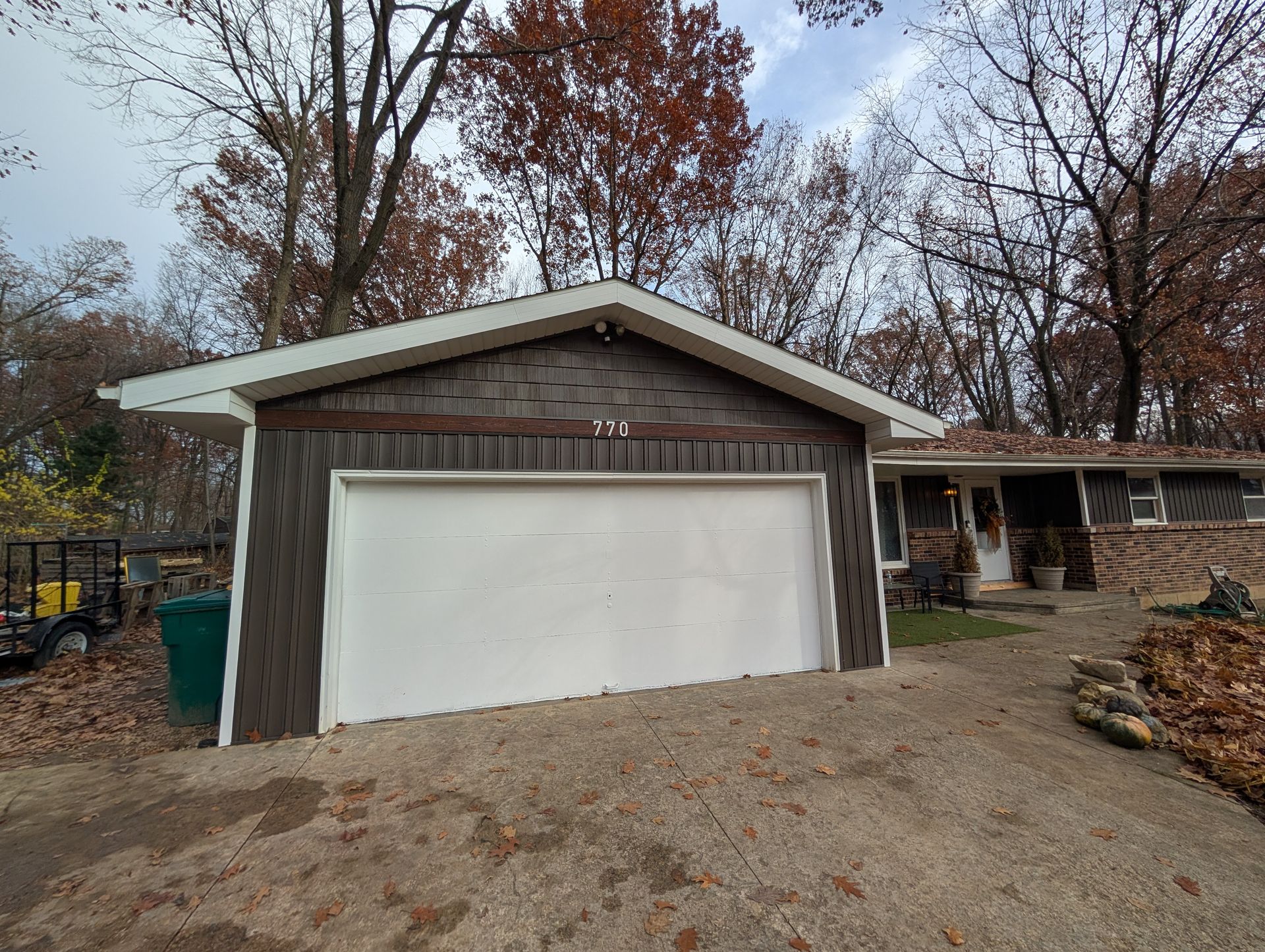 Garage with white door, brown siding, and light-colored roof. House visible to the right, surrounded by trees.
