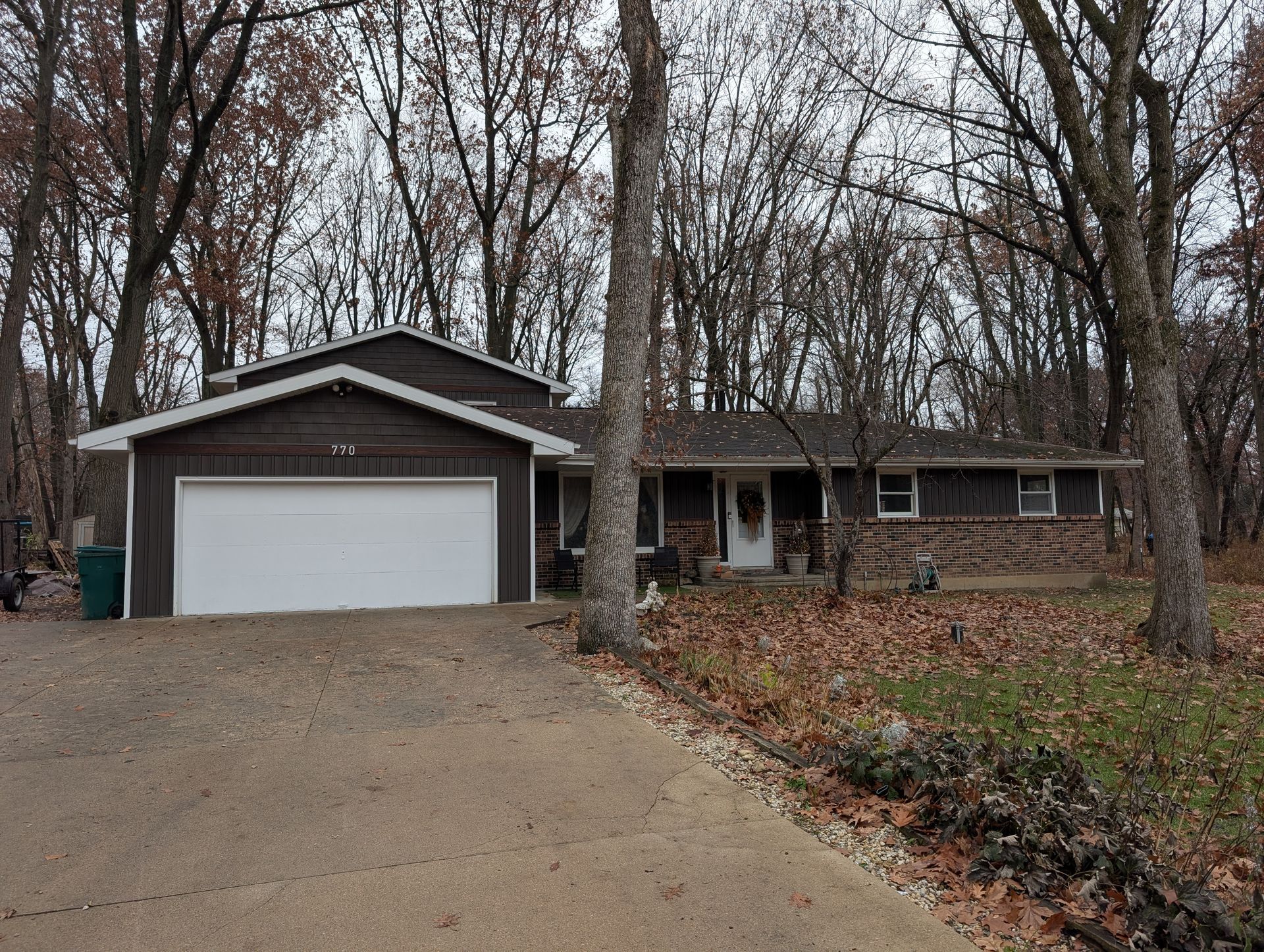 Garage with light yellow siding, closed door, and white table outside on a concrete driveway; car parked next to it.