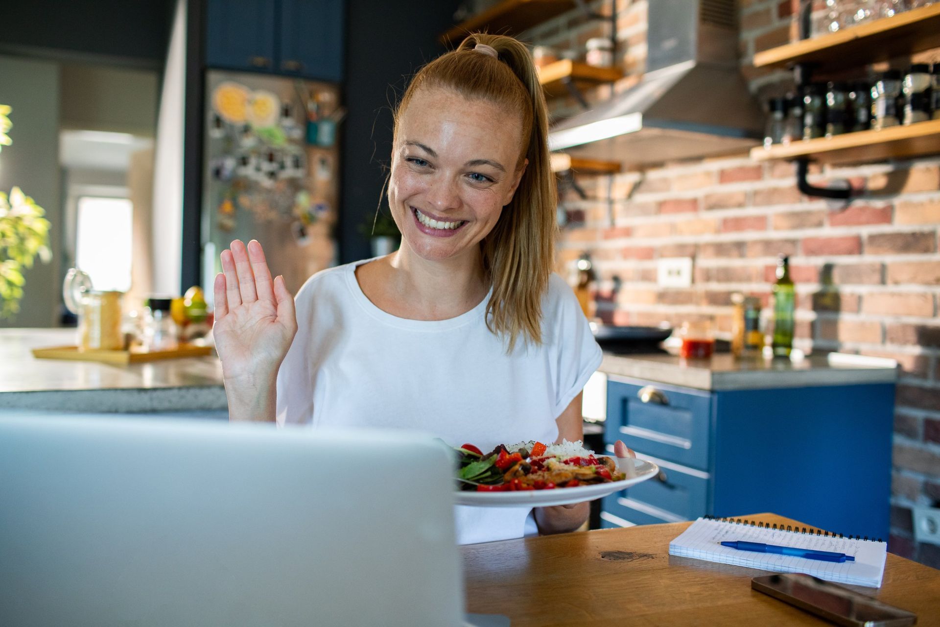 Smiling woman waves on a video call at a laptop, seated at a kitchen table with a plate of salad.